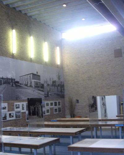 The interior of a museum exhibition space featuring brick walls, vertical light fixtures, display tables, and large black-and-white historical photographs mounted on the walls.