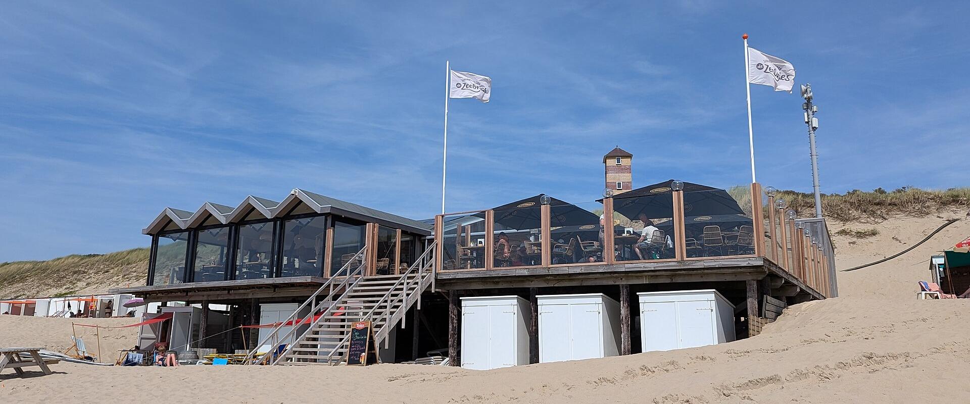 A wooden beach pavilion on stilts with a raised deck, glass-enclosed seating area, and two white flags bearing the logo "Zeebries" flying above it, set against sand dunes and a blue sky.