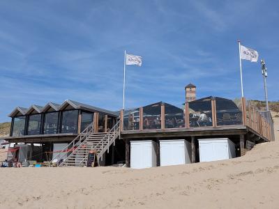 A wooden beach pavilion on stilts with a raised deck, glass-enclosed seating area, and two white flags bearing the logo "Zeebries" flying above it, set against sand dunes and a blue sky.