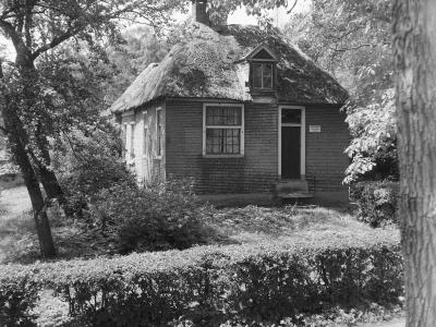 A black and white photograph of a small, traditional brick cottage with a thatched roof, surrounded by trees and a hedge.