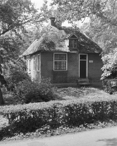 A black and white photograph of a small, traditional brick cottage with a thatched roof, surrounded by trees and a hedge.