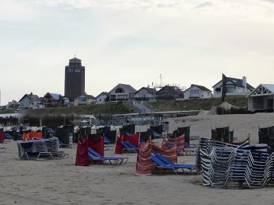 A sandy beach with stacks of blue lounge chairs and red privacy screens in the foreground, backed by dunes and a row of beach houses, with a prominent dark brick tower visible in the background.