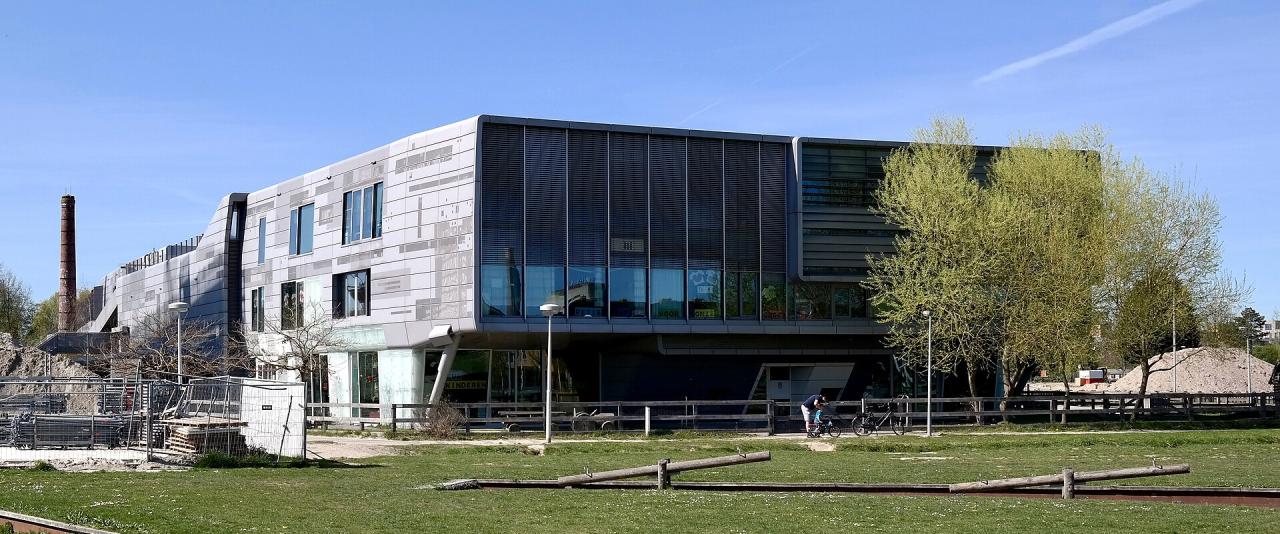 1. A modern, angular building with a grey tiled facade and large glass windows, situated next to a grassy area with wooden logs and a brick chimney in the background.