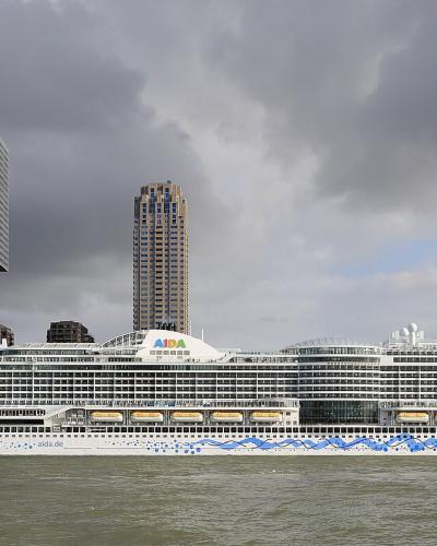 A large white cruise ship with a distinctive blue wave pattern and a giant eye painted on its bow is docked at a port, with a backdrop of modern high-rise buildings including the De Rotterdam complex and the Erasmus Bridge area under a cloudy sky.