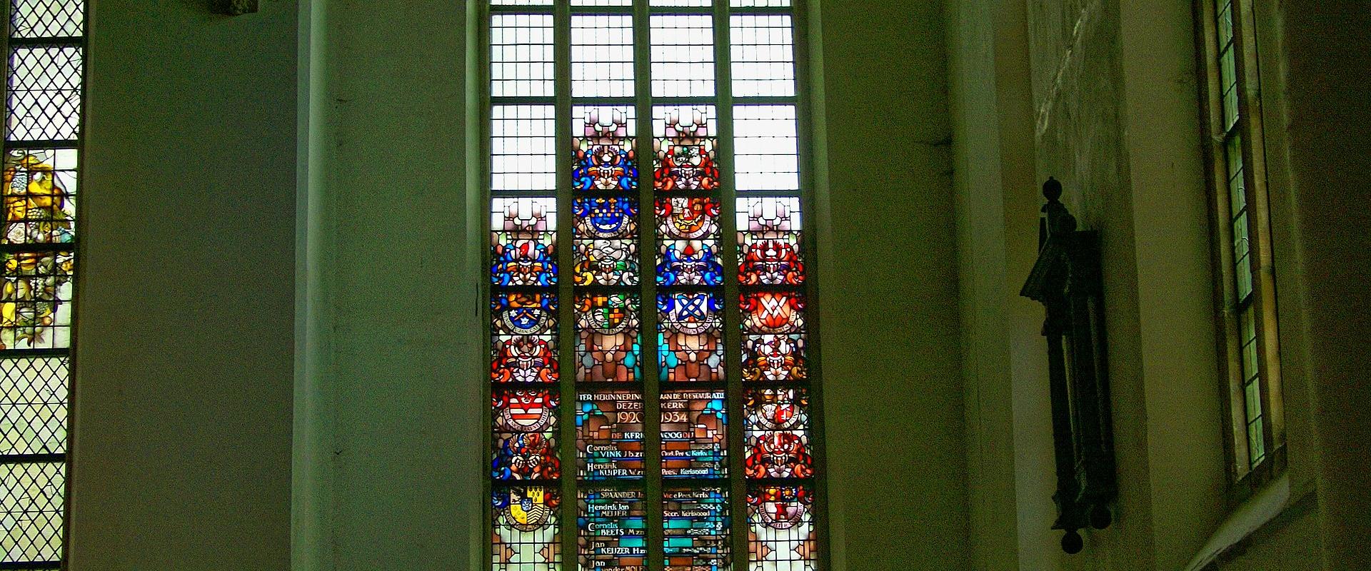 Interior view of a large church hall featuring tall Gothic arched windows with stained glass, a dark wooden ceiling, and a shop area at the bottom with a sign reading "WERELDWINKEL".