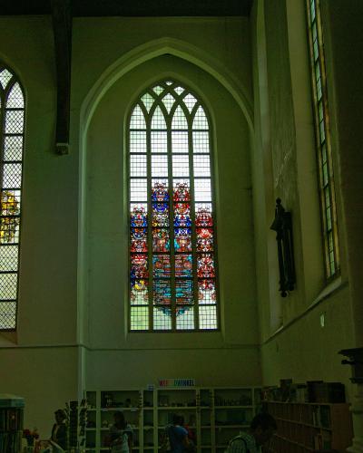 Interior view of a large church hall featuring tall Gothic arched windows with stained glass, a dark wooden ceiling, and a shop area at the bottom with a sign reading "WERELDWINKEL".