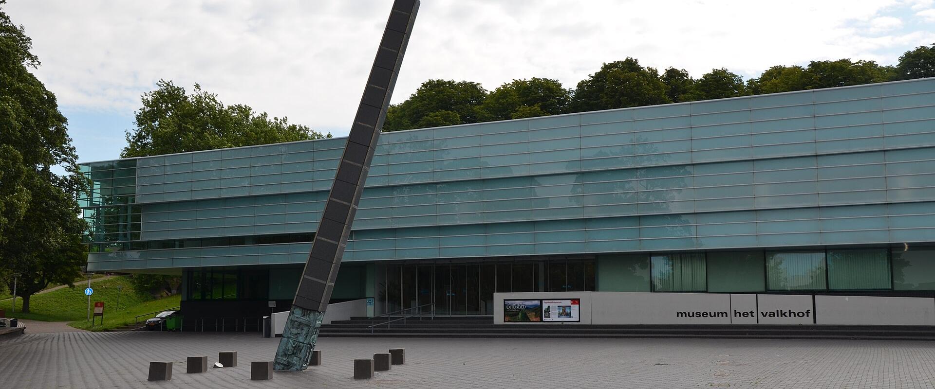 A modern glass-fronted building with a large leaning sculpture in front, featuring signage that reads "museum het valkhof".