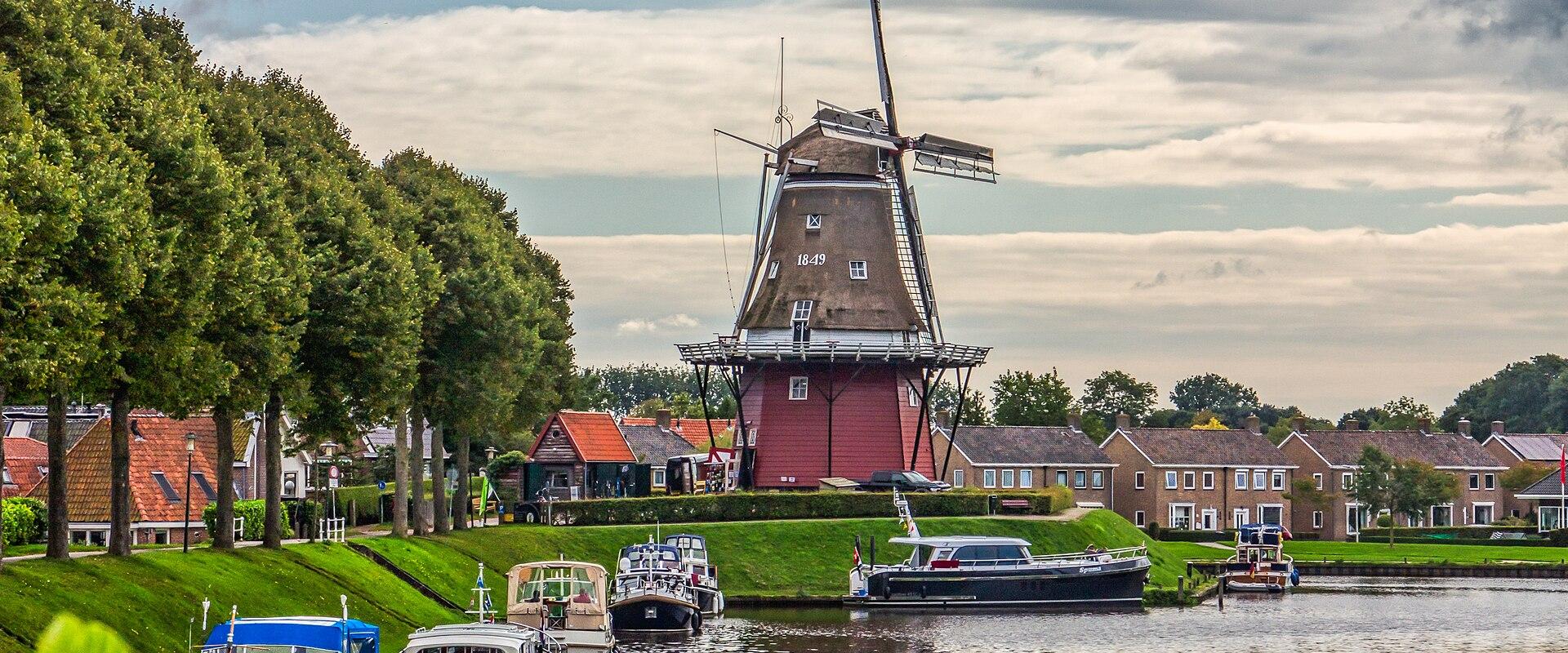 A red smock windmill stands on a grassy bank next to a canal with moored boats, framed by trees and houses under a cloudy sky.