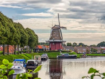 A red smock windmill stands on a grassy bank next to a canal with moored boats, framed by trees and houses under a cloudy sky.