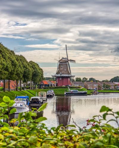 A red smock windmill stands on a grassy bank next to a canal with moored boats, framed by trees and houses under a cloudy sky.