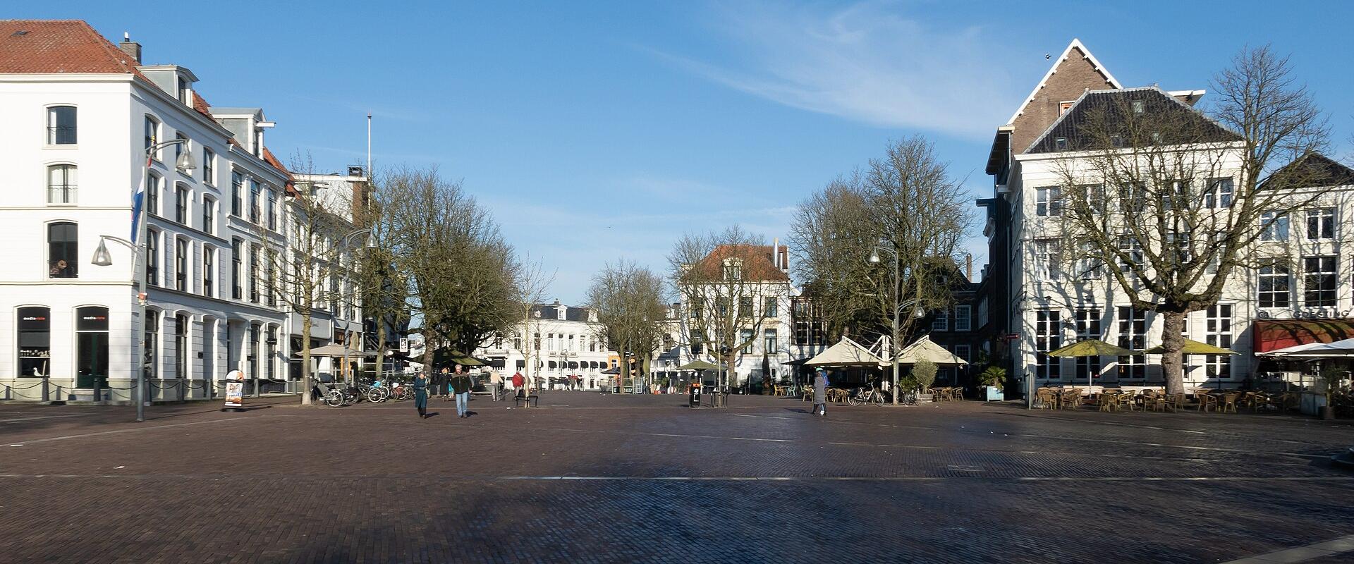 A wide, paved public square (brink) lined with historic Dutch buildings, trees, and outdoor cafe seating under a blue sky.