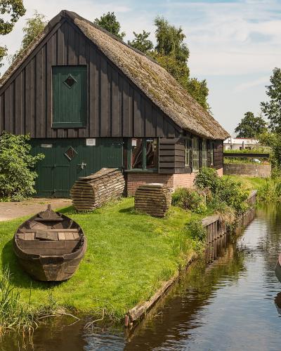 A narrow canal lined with moored boats and traditional thatched-roof wooden buildings, with stacks of wooden planks on the grassy bank.