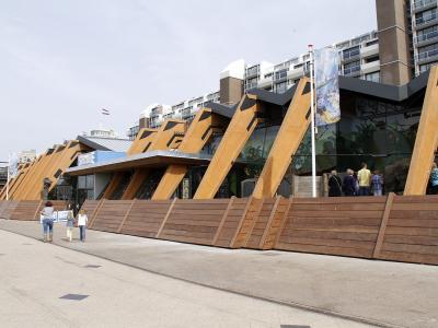 The image shows a paved promenade next to a body of water, featuring a large, dark, angular architectural structure on the left, a row of tall streetlights, and a whimsical metal sculpture of a figure in the distance.