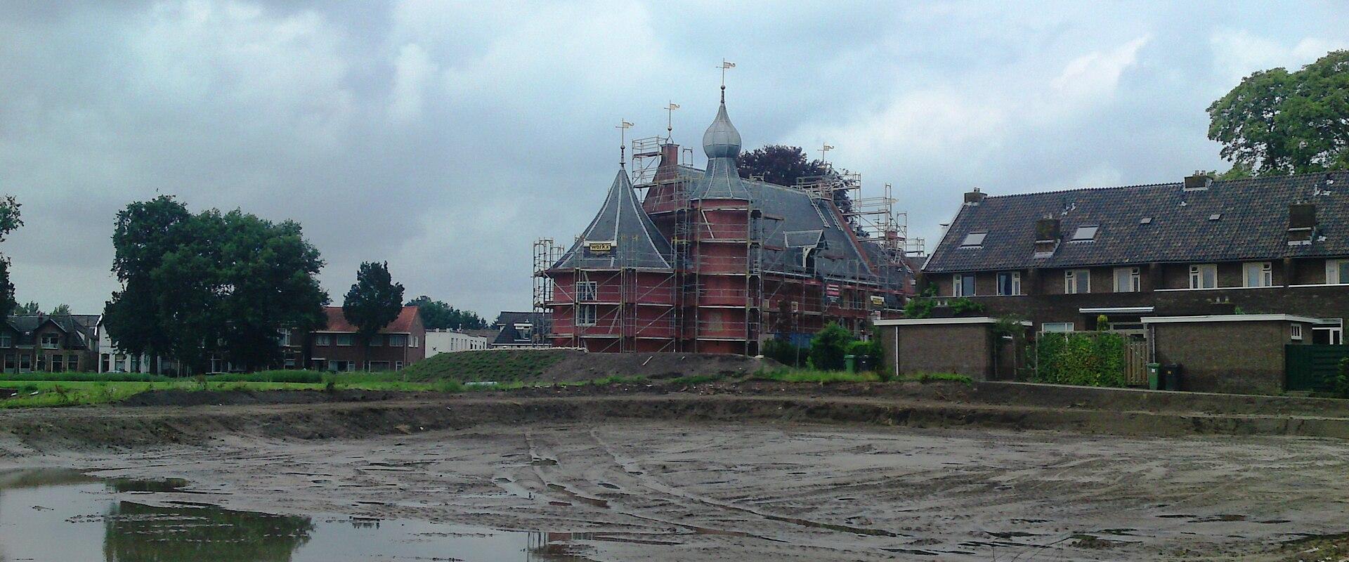 A large red-brick castle with multiple turrets and a central spire, currently covered in scaffolding for renovation, situated behind a wide, muddy dry moat or construction site.