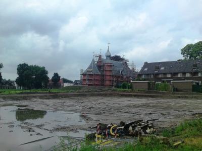 A large red-brick castle with multiple turrets and a central spire, currently covered in scaffolding for renovation, situated behind a wide, muddy dry moat or construction site.