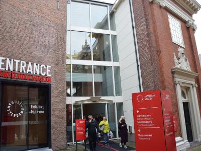 1. The image shows the entrance to a building with prominent signage reading "ENTRANCE AMSTERDAM MUSEUM" in white and red letters on a brick facade, alongside a modern glass extension and a red directional sign listing "Amsterdam Museum," "Schuttersg