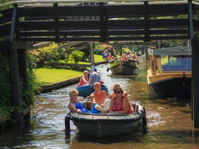 A group of tourists in small electric boats navigating a narrow canal under a wooden footbridge, with other boats and greenery visible in the background.