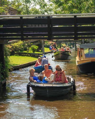A group of tourists in small electric boats navigating a narrow canal under a wooden footbridge, with other boats and greenery visible in the background.
