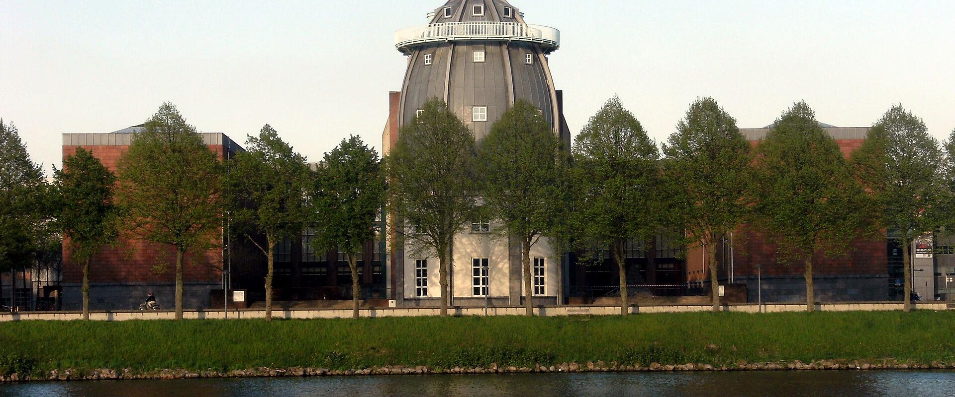 A large, distinctive building with a prominent grey dome and brick wings, viewed from across a river with a line of trees in the foreground.