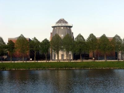 A large, distinctive building with a prominent grey dome and brick wings, viewed from across a river with a line of trees in the foreground.