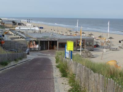 A paved path leads to a beachside building with outdoor seating, set against a backdrop of sand dunes and the sea.