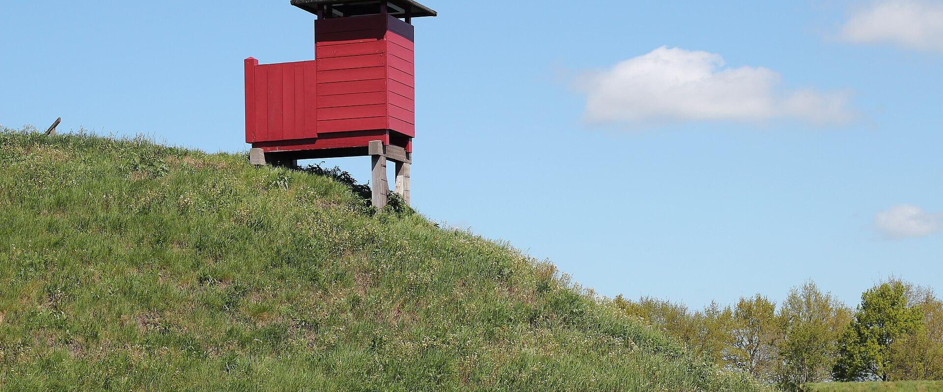 A red wooden watchtower or guard post situated on top of a grassy earthen rampart under a blue sky.