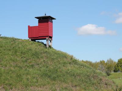 A red wooden watchtower or guard post situated on top of a grassy earthen rampart under a blue sky.
