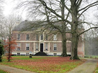 A large, symmetrical brick manor house with white window frames and a central entrance, viewed through bare trees in a park-like setting with fallen autumn leaves.