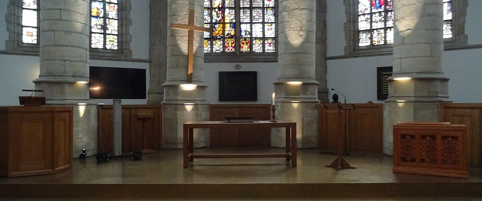 The image shows the interior of a large church featuring massive cylindrical stone pillars, tall stained-glass windows, and a wooden altar area with a cross.